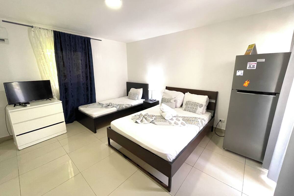 Een hotelkamer met twee eenpersoonsbedden, een groot bed, een televisie op een dressoir, een koelkast en een raam met gordijnen.