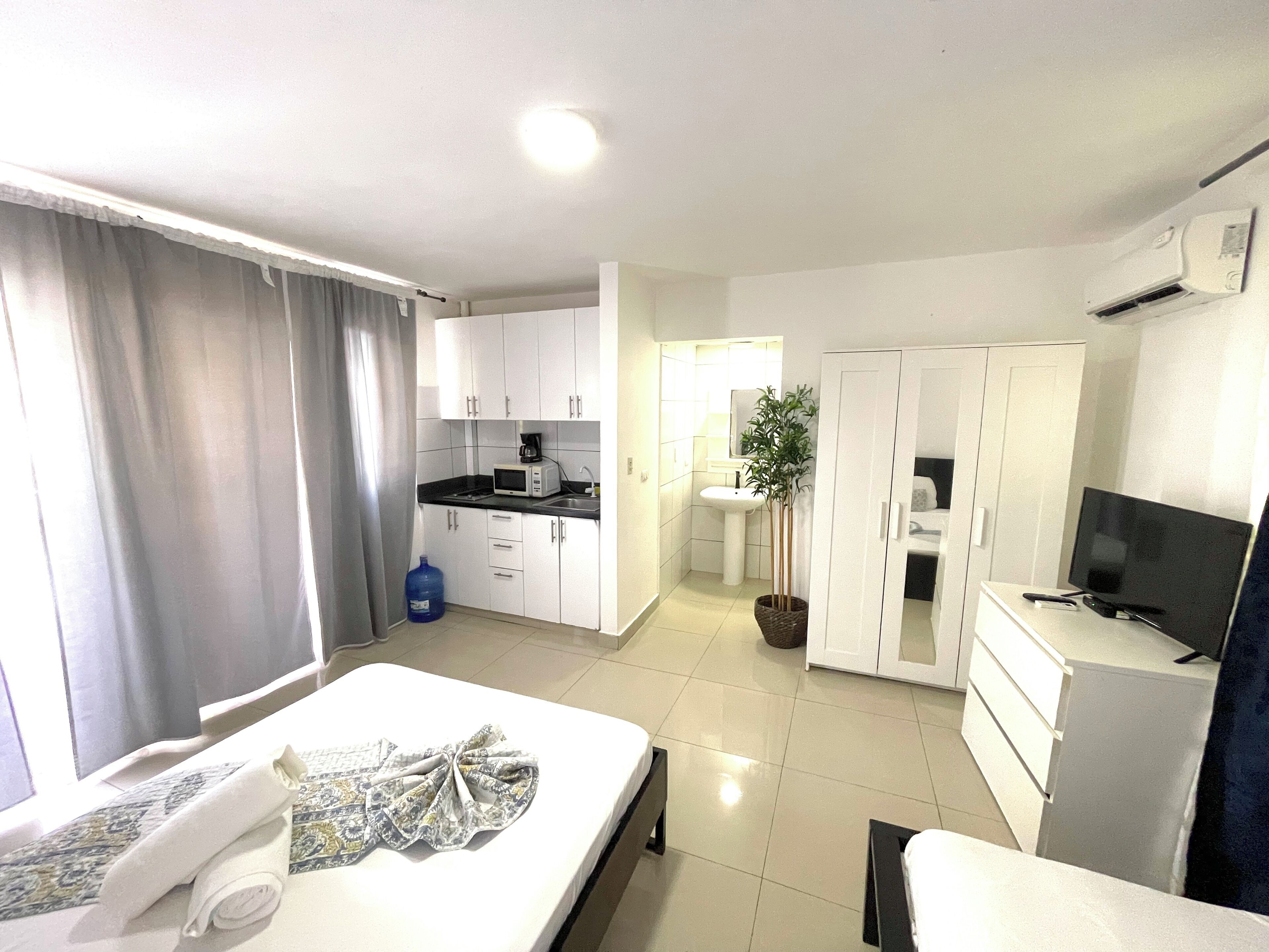 A compact hotel room with a bed, a small kitchenette, a bathroom, and a TV.
