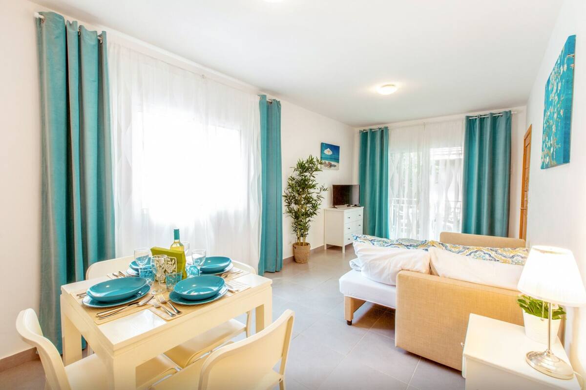 Een goed verlichte kamer met een eettafel gedekt voor twee personen, een bed met een wit- en blauw gebed eiland, en een televisie op een witte dressoir.