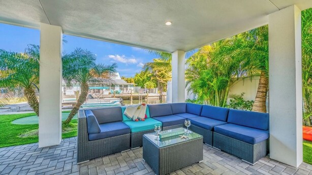 A covered outdoor seating area with a large sectional sofa, a glass-top coffee table, and a view of a pool and greenery. - Amazing home on canal! Paddle boards, and kayaks! (Bradenton Beach)
