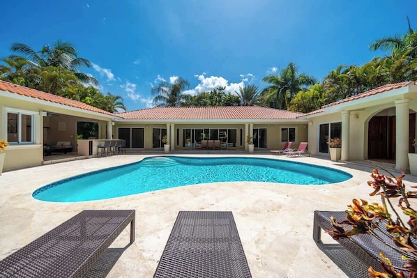 Une piscine avec des chaises longues, un patio couvert et des palmiers.