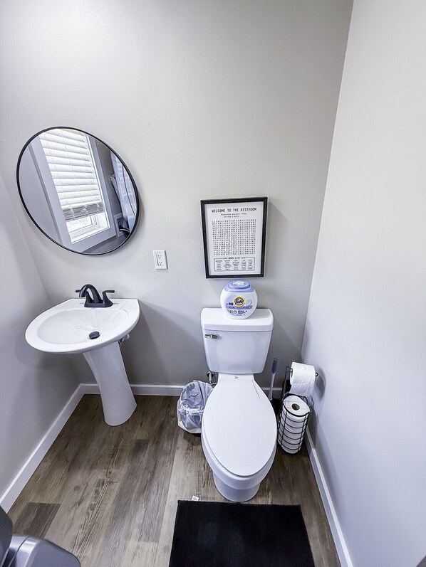 Et badeværelse med en hvid vask, toilet, et indrammet ordgætteleg og et spejl.