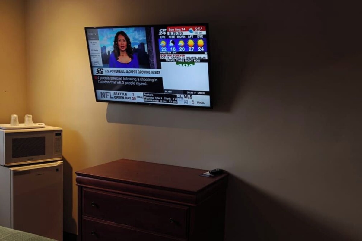 A hotel room with a television mounted on the wall displaying a news channel, a microwave, a wooden dresser, and a bed with a green bedspread.