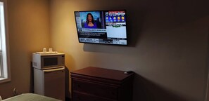 A hotel room with a television mounted on the wall displaying a news channel, a microwave, a wooden dresser, and a bed with a green bedspread. - Queensway Motel (Toronto)