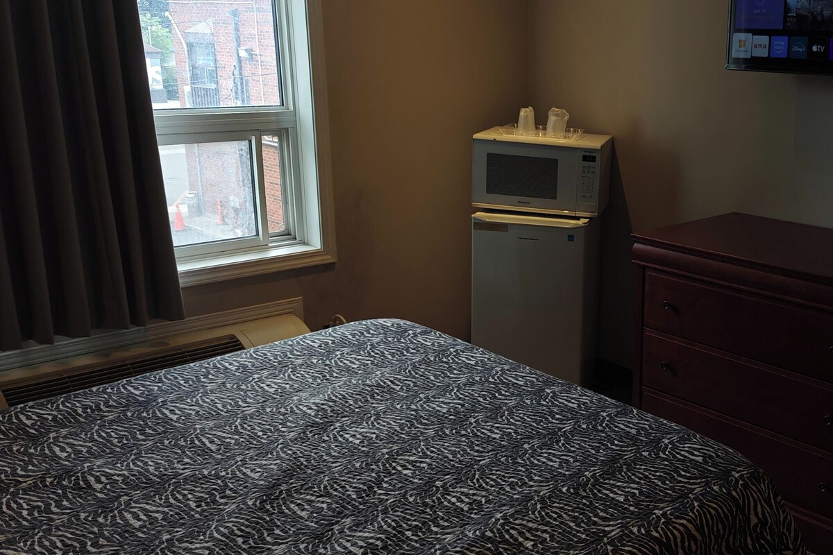 A bedroom with a patterned bed, a small refrigerator, a TV mounted on the wall, and a window with curtains.
