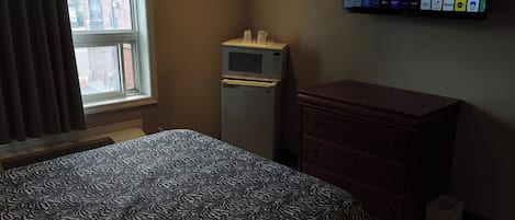 A hotel room with a bed, a small refrigerator, a wooden dresser, and a window with curtains.
