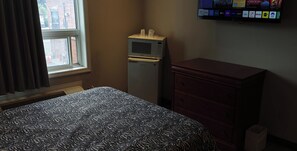 A hotel room with a bed, a small refrigerator, a wooden dresser, and a window with curtains. - Queensway Motel (Toronto)