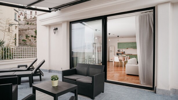 Un balcon doté de meubles en osier, d’une plante en pot et offrant une vue sur la cuisine et le salon, visibles à travers une porte vitrée.