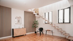 Un intérieur moderne avec un parquet en bois disposé en chevrons, une chaise noire, une petite table, un meuble en bois, un miroir, un escalier et une fenêtre.