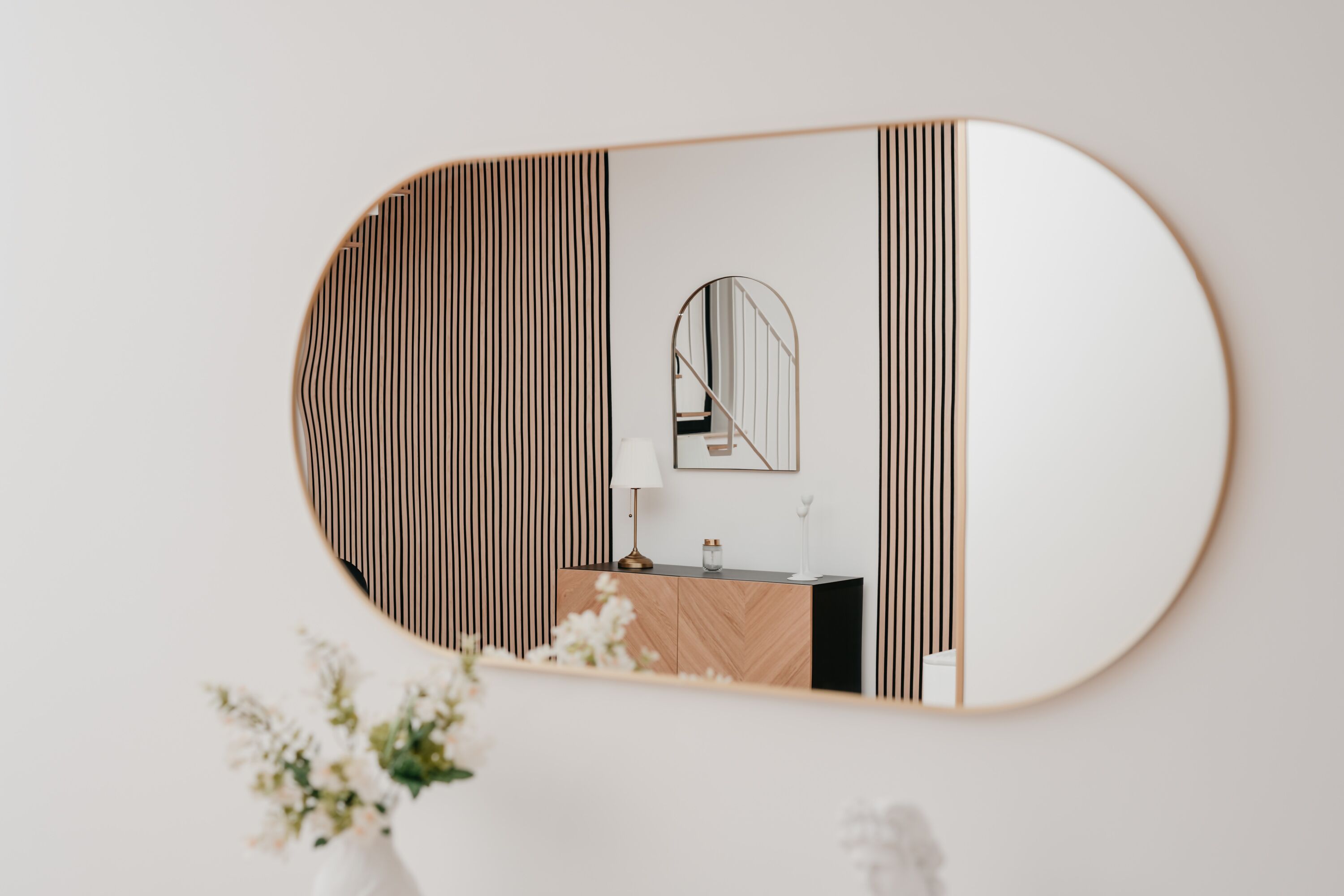 Un espejo con marco de madera que refleja una habitación con pared de diseño espiga, un espejo redondo y un mueble de madera con una lámpara.