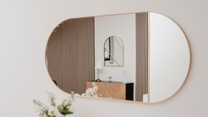 Un miroir avec un cadre en bois reflétant une pièce dont le mur est décoré d’un motif en chevrons, un miroir rond et un meuble en bois sur lequel est posée une lampe.