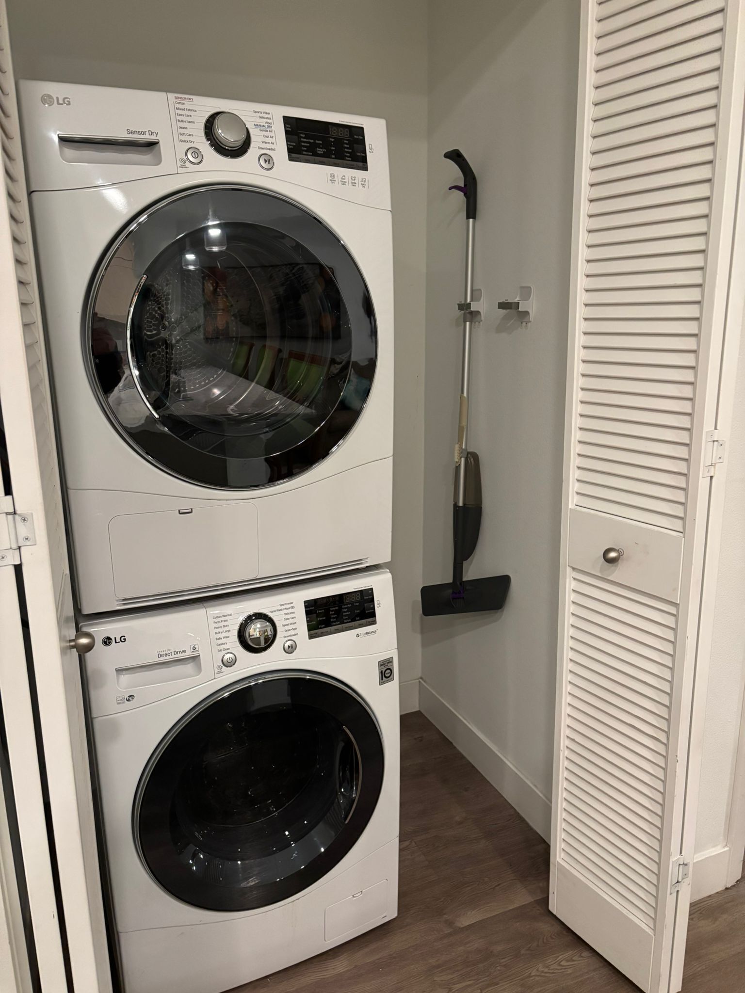 A stacked washer and dryer unit with a broom leaning against the wall.