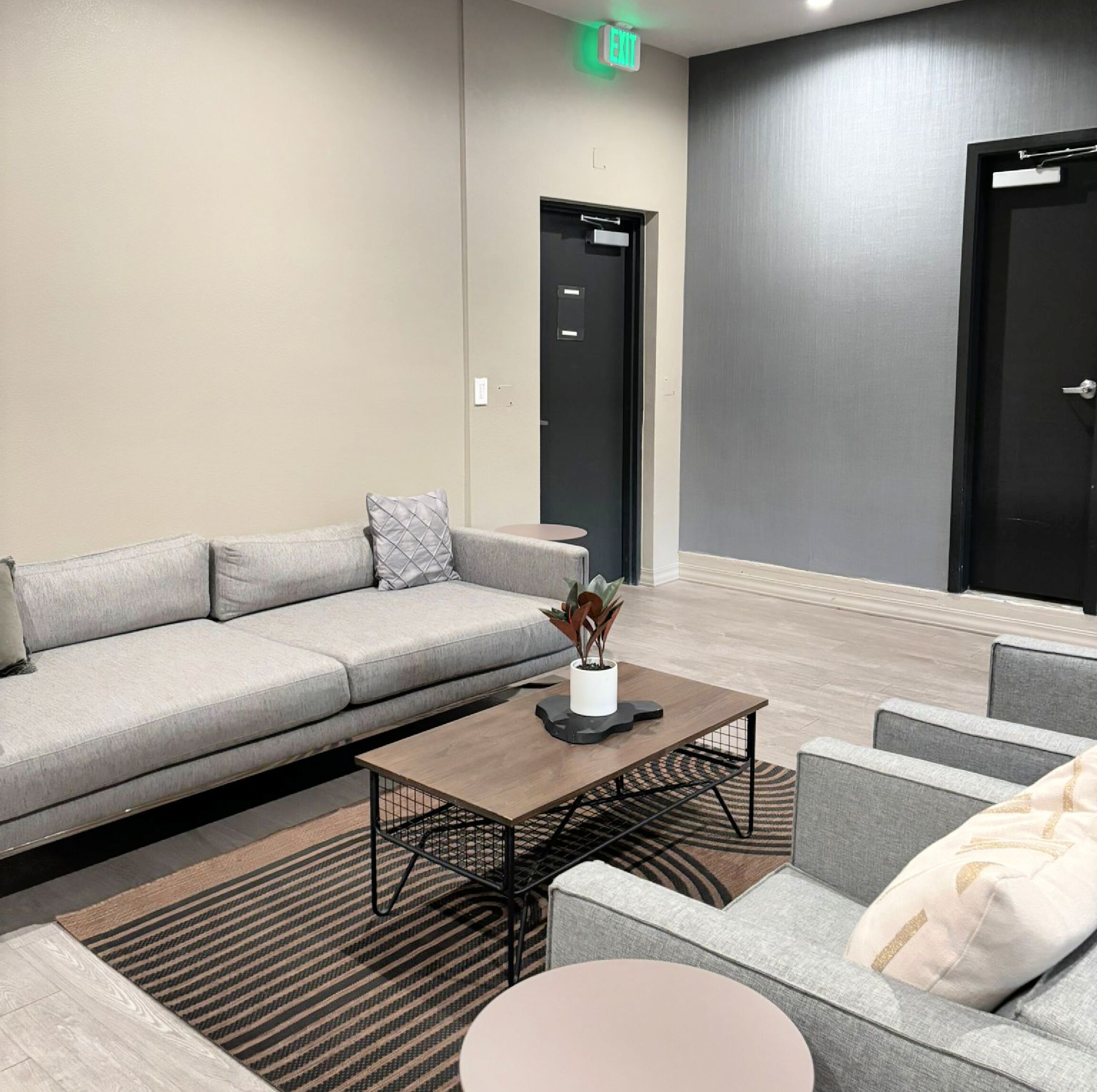 Un hall d’entrée moderne avec un canapé gris, une table basse et une petite table sur laquelle est posée une plante. On aperçoit deux portes au fond, dont l’une porte un panneau indiquant la sortie.