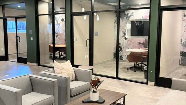 A modern office space with grey sofas, a coffee table, and a meeting room visible through glass doors.