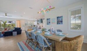 Un comedor moderno con una larga mesa dispuesta para cuatro personas, rodeada de sillas de mimbre, una cocina al fondo y un balcón con vistas.