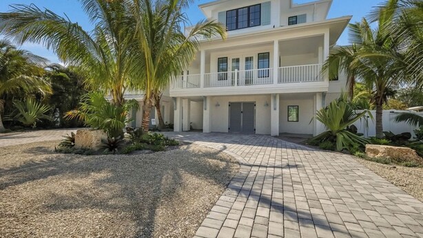 A two-story building with a balcony, surrounded by palm trees and a gravel driveway. - Thanksgiving availability! Stunning pool home! (Holmes Beach)