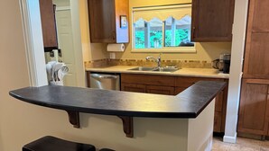 A kitchen with a central island, two bar stools, wooden cabinets, a window with a view, and a built-in dishwasher.