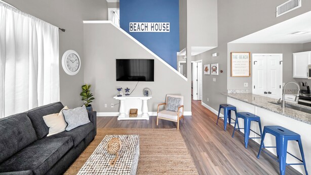 A modern living room with a grey sofa, a coffee table, and a TV. A staircase leads to a kitchen with blue bar stools. - SandSational Staycation Pet Friendly, Fenced Yard, 6min to the Beach (Navarre Beach)