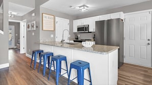 A modern kitchen with white cabinets, a stainless steel refrigerator, a microwave, and a granite countertop. There are three blue bar stools and a framed quote on the wall. - SandSational Staycation Pet Friendly, Fenced Yard, 6min to the Beach (Navarre Beach)