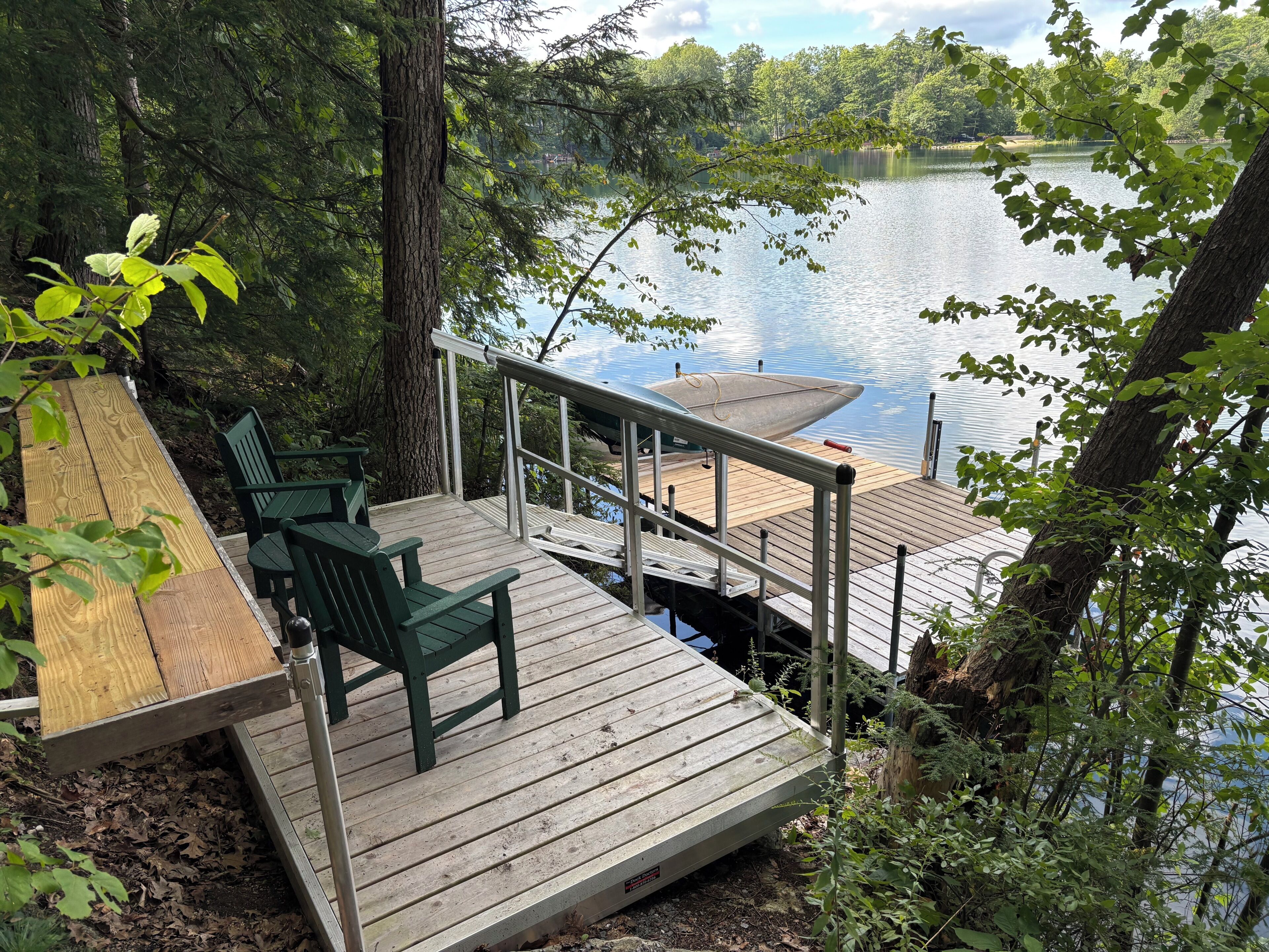 Une terrasse en bois avec des chaises, donnant sur un lac paisible, le tout entouré d’arbres.