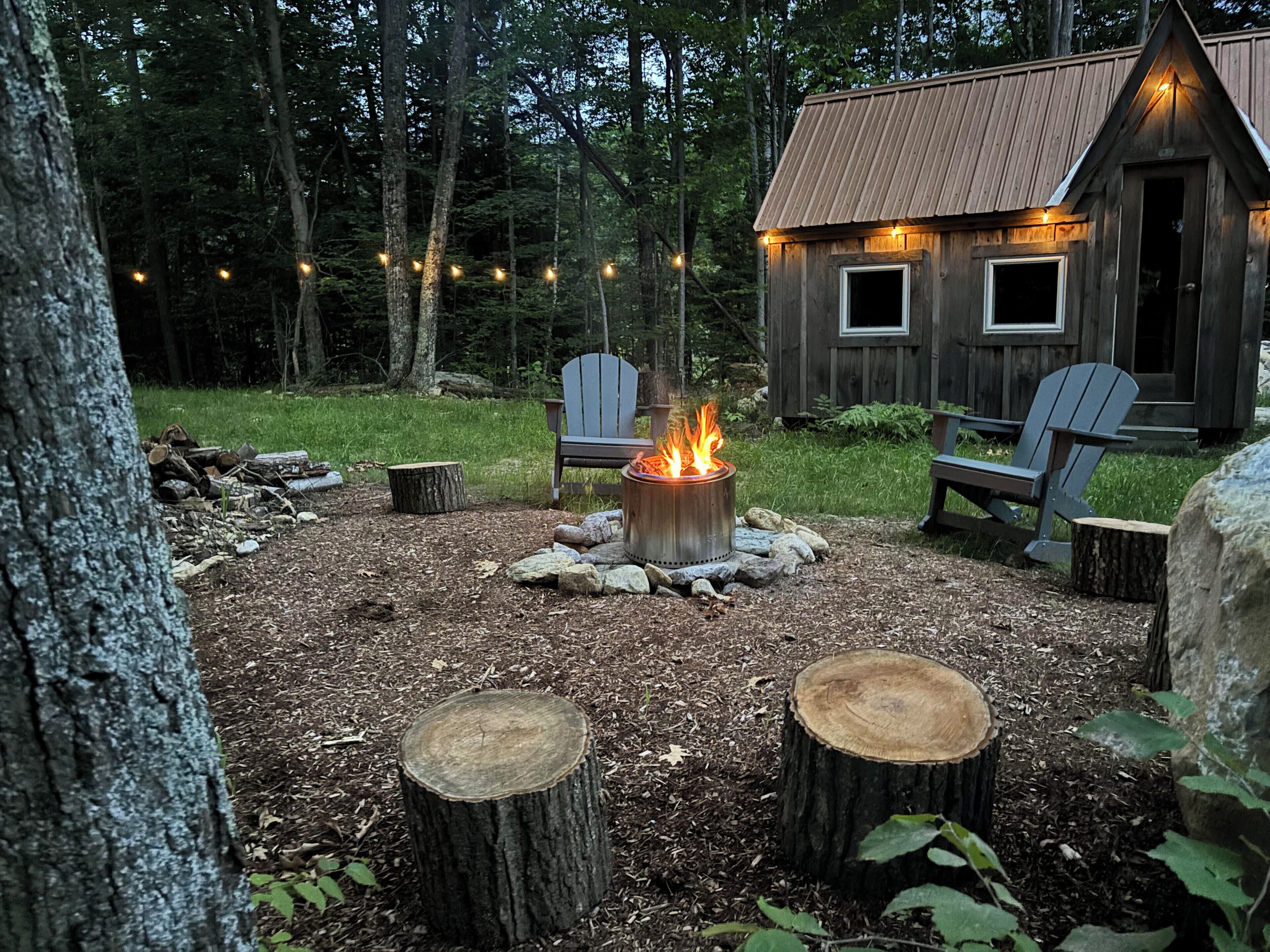 Eine rustikale Holzhütte mit einem Feuerkorb, umgeben von Adirondack-Stühlen, inmitten eines bewaldeten Gebietes.