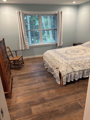 A bedroom with a wooden floor, a bed with a floral bedspread, a dresser, a rocking chair, and a window with curtains.