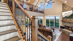 A spacious living area with a wooden staircase, a fireplace, and a large window offering a scenic view. - Stunning Views | Sauna | Hot tub | Bar- 2 Kitchens (Ranger)