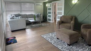 Ein Wohnzimmer mit grauem Sofa, braunem Sessel und Couchtisch. Auf dem Boden liegt ein Teppich mit Muster, und im Hintergrund ist eine Tür mit satinierten Glasscheiben zu sehen.