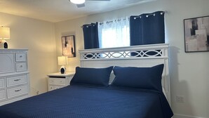 Ein Schlafzimmer mit Bett, Nachttisch, Lampe, Deckenventilator und einem Fenster mit Vorhängen.