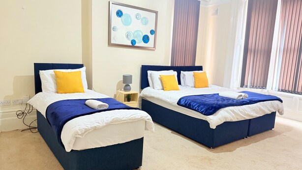 A hotel room with two beds, each with a blue headboard, white bedding, and yellow pillows. There is a painting on the wall and a window with curtains. - 1 Bed Flat - Sleeps 5 - Parking - Wifi (Leicester)