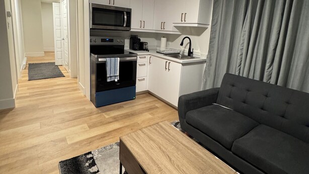 Un salon moderne avec un canapé foncé, une table basse en bois et un coin cuisine doté de meubles blancs et d’un micro-ondes noir.