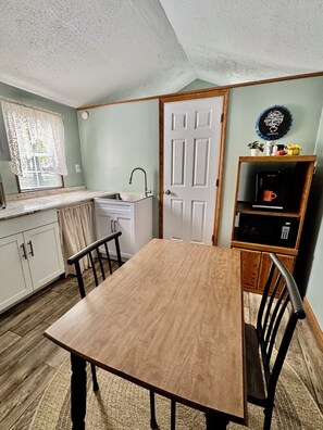 A compact kitchen with a sink, a window with blinds, a door, and a small shelf with a coffee maker and decorative items. - Corbin Cottage | Near Corbin Arena, Laurel River Lake & Cumberland Falls (Corbin)
