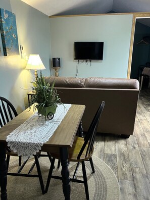 A dining area with a table, chairs, a lamp, a plant, a sofa, and a TV mounted on the wall.