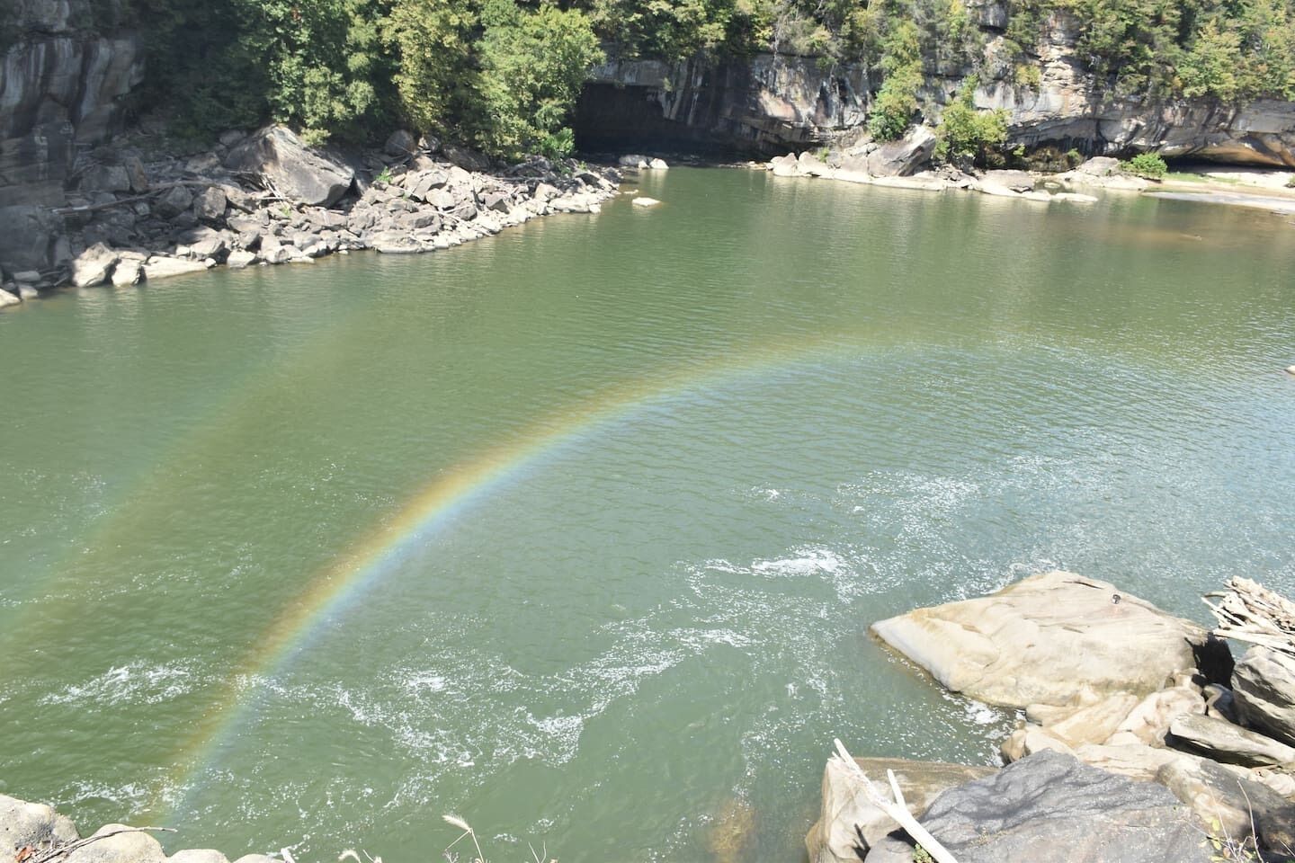 A river with a visible rainbow, surrounded by rocky terrain and greenery.