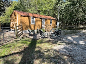 A small, single-story building with a brown exterior and a red roof, surrounded by trees and a gravel area with outdoor furniture. - Corbin Cottage | Near Corbin Arena, Laurel River Lake & Cumberland Falls (Corbin)