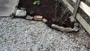 Ein Blumenbeet mit Pflanzen, ein Kiesweg und ein Holenzaun.