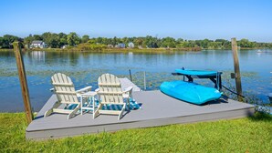 Een steiger met twee Adirondack stoelen en een blauwe opblaasbare luchtbed op een gazon aan een rustig meer, omringd door bomen en huizen op de achtergrond.