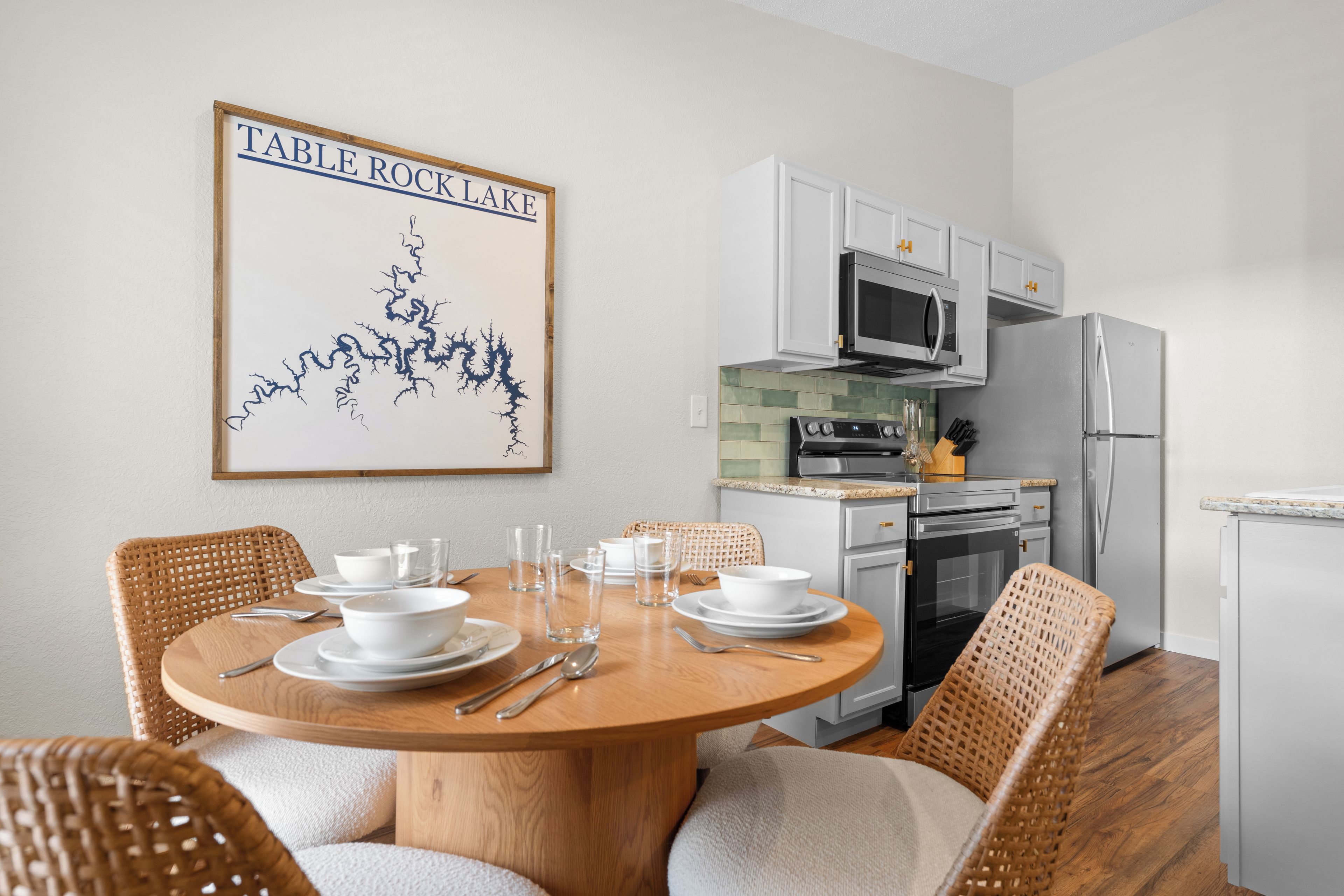 A dining area with a round wooden table set for four, surrounded by wicker chairs. A kitchen with white cabinets and stainless steel appliances is visible in the background.