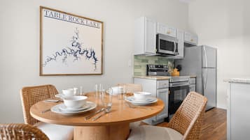 A dining area with a round wooden table set for four, surrounded by wicker chairs. A kitchen with white cabinets and stainless steel appliances is visible in the background.