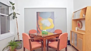 Un comedor con una mesa de madera y cuatro sillas anaranjadas, un cuadro abstracto grande en la pared, y un mueble de madera con un reloj y adornos.
