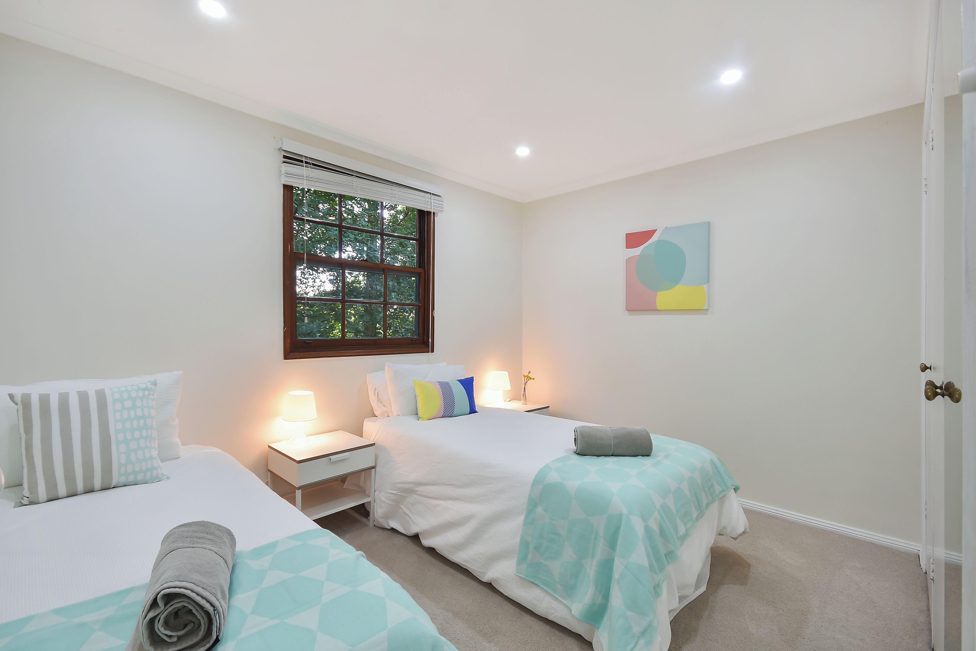 A bedroom with two beds, a window with blinds, a bedside table with a lamp, and a wall-mounted artwork.