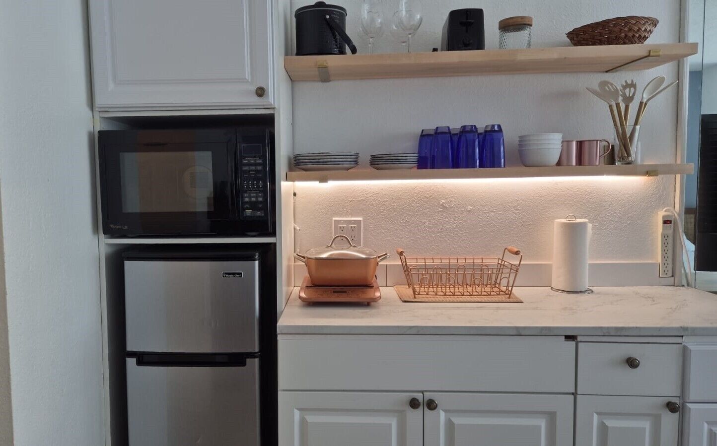 Una cucina con forno a microonde, frigorifero, una pentola sul piano di lavoro e scaffali con utensili da cucina.