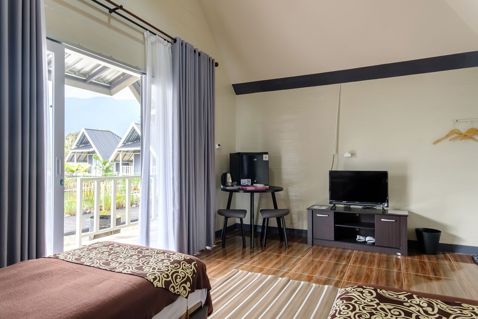 Habitación de hotel con cama, televisión, escritorio y balcón con vistas a casas.