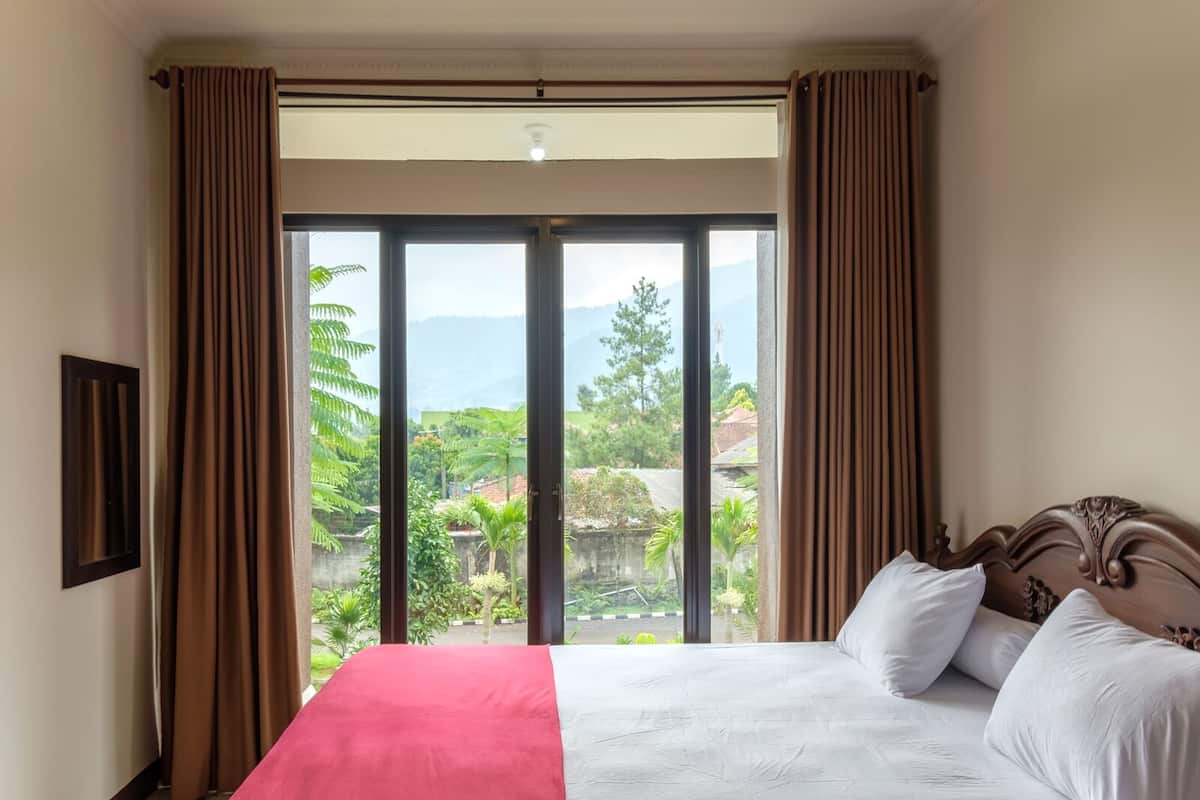 Quarto com cama, cortinas e vista para o verde e montanhas.