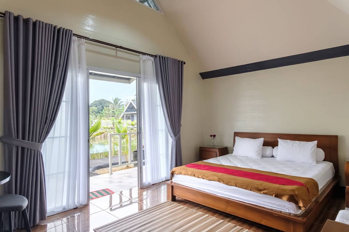 Quarto com cama, mesa de cabeceira, uma cadeira e vista para uma piscina e vegetação lá fora.