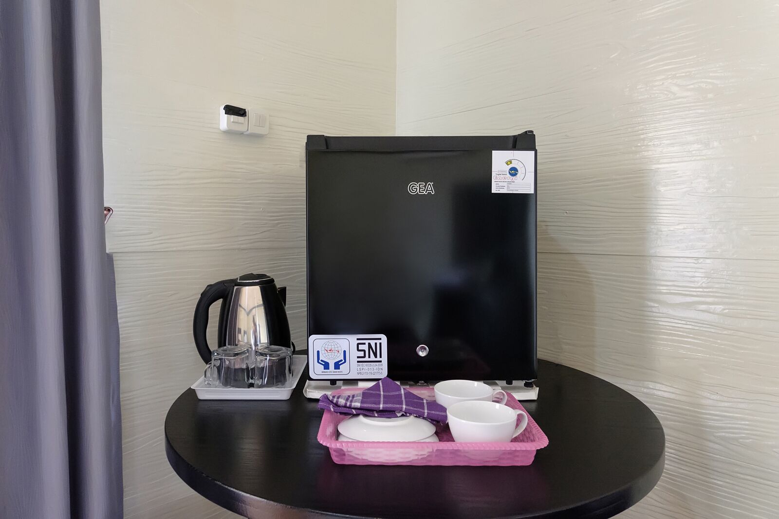 A GEA refrigerator with a SNI certification label, a kettle, a tray with cups and a plate, and a power outlet on the wall.