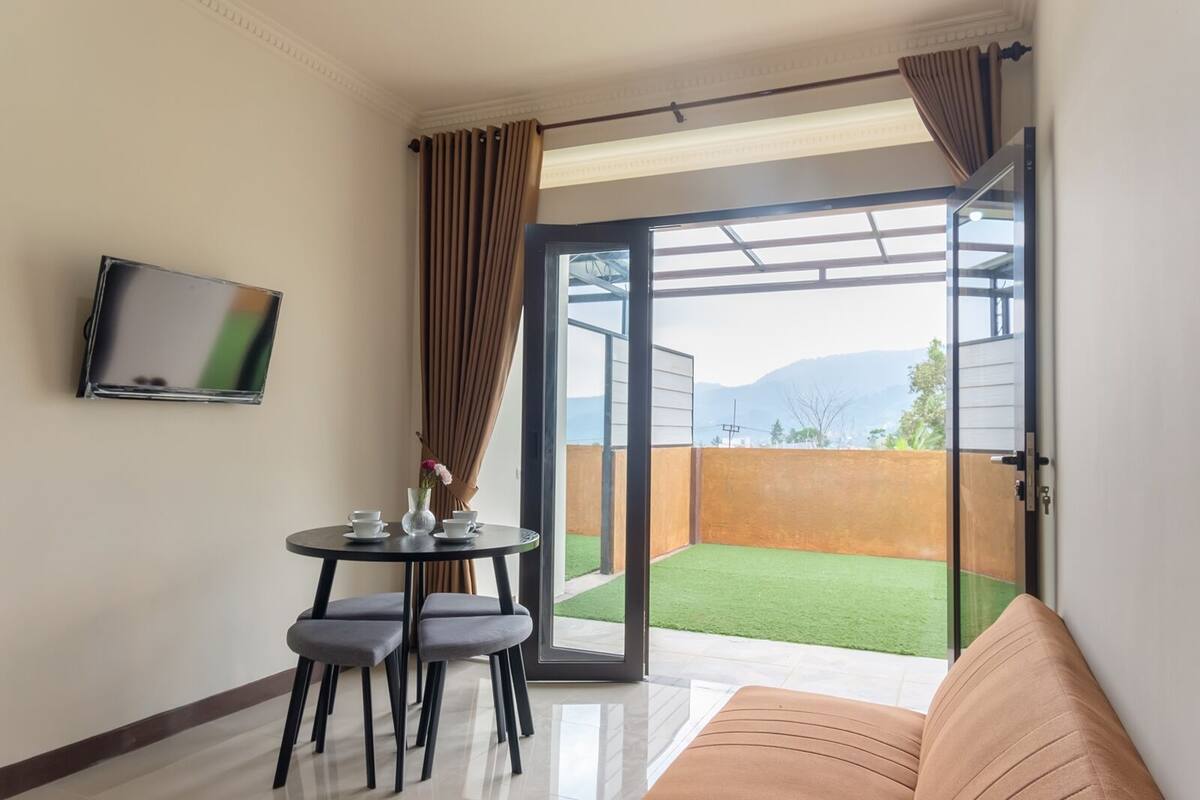 Quarto moderno com sofá, mesa com xícaras e um vaso, TV na parede e porta de vidro deslizante que dá para uma varanda com vista para as montanhas.
