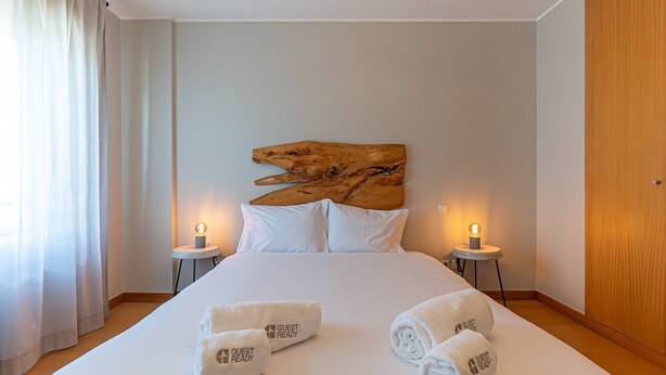 A hotel room with a large bed, two bedside lamps, a wooden headboard, and rolled towels with "GUEST READY" text. - GuestReady - Serenity home in Porto (Boavista)