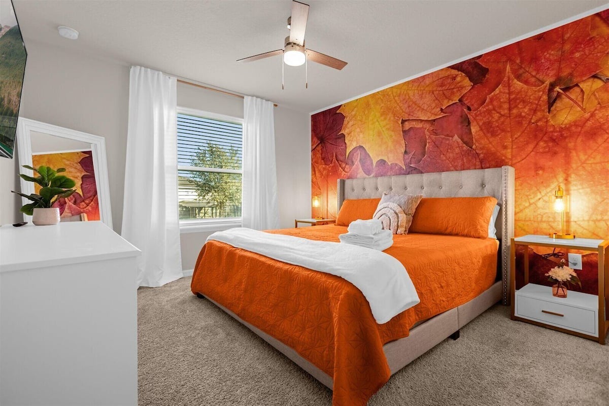 A hotel room with a large bed, a ceiling fan, a window with white curtains, and a wall mural.