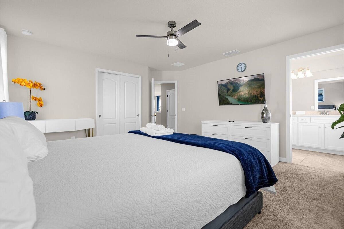 A bedroom with a large bed, a ceiling fan, a painting, and a bathroom with a vanity and mirror.